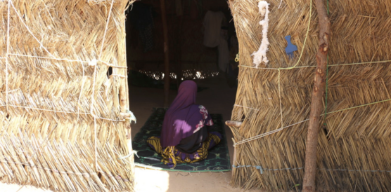 Das Bild zeigt eine Hütte aus Stroh, im Eingang sitzt ein Mädchen, den Rücken zur Kamera gewandt 