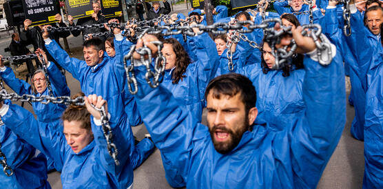 Das Bild zeigt eine Protestaktion, mehrere Menschen mit blauen Overalls knien auf dem Boden und halten schwere Eisenketten in de Höhe