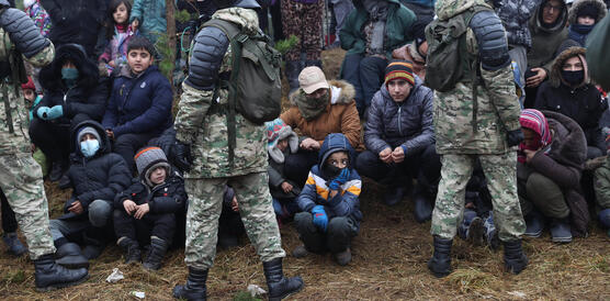 Das Bild zeigt Soldaten im Vordergrund, im Hintergrund sitzen Kinder und Erwachsene auf dem Boden