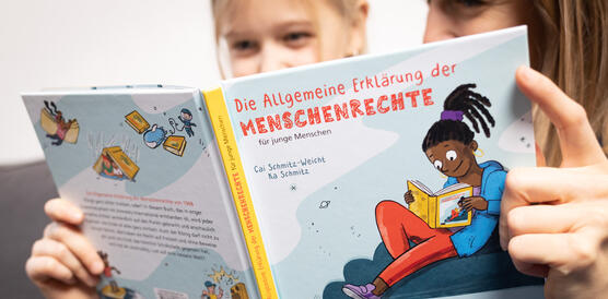 Das Bild zeigt eine junges Mädchen und Frau, die gemeinsam ein Buch halten. Darauf steht "Allgemeine Erklärung der Menschenrechte für junge Menschen". 