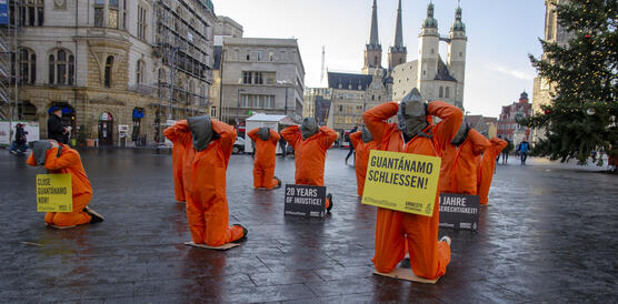 Das Bild zeigt Demonstrierende in orangenen Overalls mit Säcken über den Köpfen, die mit ihren Armen hinter dem Kopf auf dem Boden knien. Manche der Menschen tragen Amnesty-Schilder mit Aufschriften wie "Guantanamo schließen" um ihren Hals.