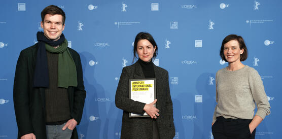 Das Bild zeigt ein Gruppenfoto mit drei Personen, eine Frau in der Mitte hält ein Dokument mit der Aufschrift "Amnesty International Film Award"