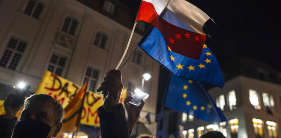 Das Bild zeigt eine Demonstration bei Nacht, die Silhouette eines Mannes, der zwei Flaggen in die Höhe hält - die EU-Flagge und die polnische Flagge