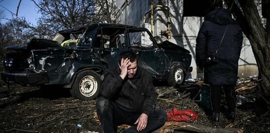Das Bild zeigt einen Mann der am Boden kniet und seinen Kopf mit der Hand stützt, im Hintergrund sieht man ein zerstörtes Auto und Gebäude.