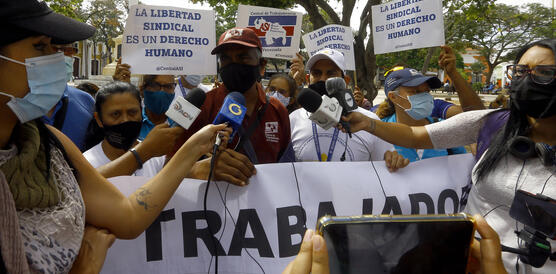 Das Bild zeigt die mehrere Menschen mit Schildern, davor stehen Reporter_innen mit Mikrofonen