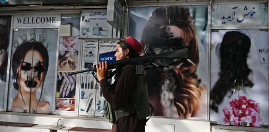 Ein Taliban-Kämpfer mit Sturmgewehr auf der Schulter geht an einem Geschäft vorbei. Auf den Plakaten des Geschäfts sind Frauengesichter schwarz übermalt.