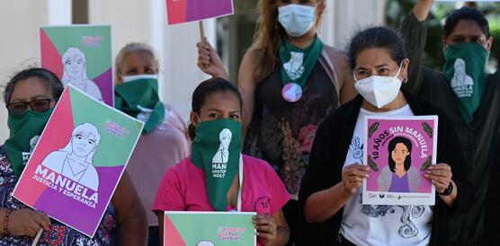 Das Foto zeigt eine Gruppe von sechs Frauen mit Mundschutz, die Schilder mit dem gezeichneten Porträt einer Frau hochhalten. Auf den Schildern steht unter anderem: Manuela.
