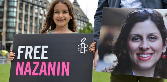 En lächelndes Mädchen hält ein Plakat in der Hand mit dem Amnesty-Logo und der Aufschrift: "Free Nazanin". Daneben steht ein Mann, der ein großes Porträtfoto von Nazanin vor sich hält.