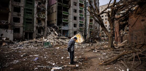 Das Bild zeigt eine Person, die vor einem stark zerstörten Haus steht