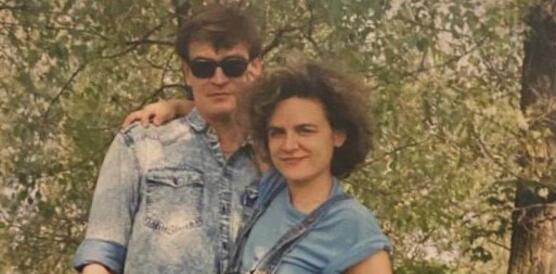 Das Bild zeigt einen Mann und eine Frau, die ihren Arm auf die Schulter des Mannes legt, sie stehen vor mehrere Bäumen, lächeln in die Kamera