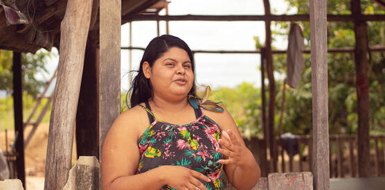 Eine indigene Aktivistin steht vor einem Holzhaus und gestikuliert beim Reden mit ihren Händen; sie trägt ein schulterfreies Kleid, ihre langen schwarzen Haare fallen über ihren Rücken.