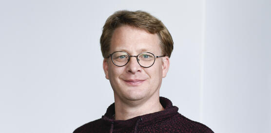 Porträtfoto von Stefan Dold, der eine Brille trägt und einen Kapuzenpulli und in die Kamera lächelt.