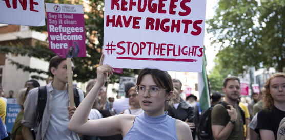 Eine Menschenmenge demonstriert auf einer Straße. Eine junge Frau im Vordergrund hält ein Schild hoch, auf dem steht: "Refugees have rights. Stop the flights".