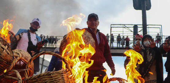 Indigene verbrennen auf einer Straße Weidenkörbe, sie stehen lichterloh in Flammen, auf einer Brücke hinter ihnen stehen Menschen.