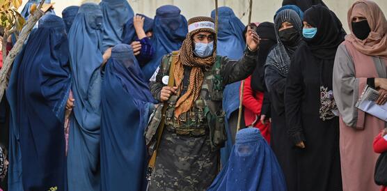 Ein mit einem Sturmgewehr bewaffneter Taliban-Kämpfer, der einen Mundschutz trägt, steht auf einem Gehweg inmitten einer Gruppe Frauen, von denen die meisten eine Burka tragen. Die anderen Frauen tragen Kopftücher und einen Mundschutz.