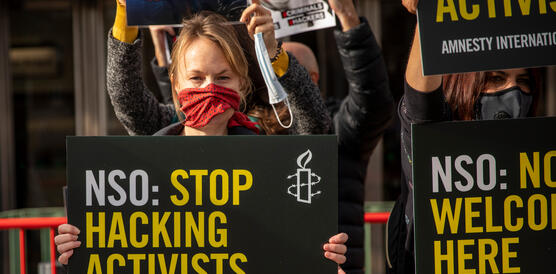 Eine Demonstration mit rotem Schal vor dem Mund hält ein Schild hoch, auf dem steht: „NSO: Stop Hacking Activists“.