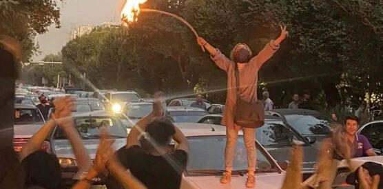 Das Bild zeigt das Porträtbild einer Frau, die mit einem brennenden Gegenstand auf einem Auto steht und die Hände in die Höhe hält.