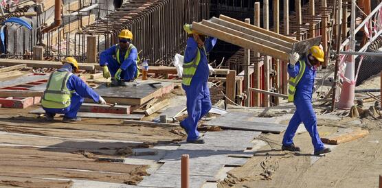 Das Bild zeigt eine Baustelle, mit mehreren Arbeitern, die gelbe Helme und Blaue Overalls tragen. 