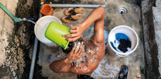 Vogelperspektive auf einen Mann, der in einer Dusche mit Steinboden steht und sich mit einem kleinen Eimer Wasser über den Kopf kippt.