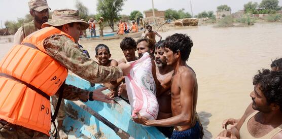 Männer in Armeekleidung und Sicherheitswesten übergeben Lebensmittel an Männer, die im Wasser stehen.