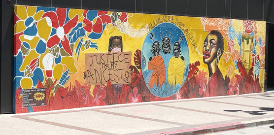 Wandbild mit floralen Mustern, einer schwarzen Frau, rituellen Masken, einem schwarzen Mädchen, das ein Schild hochhält "justice for my ancestors".