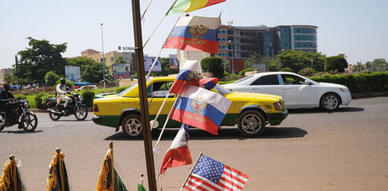 Eine Straße in Mali, auf der Autos fahren und Motorroller, am Straßenrand wehen Nationalflaggen im Wind, die US-amerikanische, die russische,die polnische, die malische.