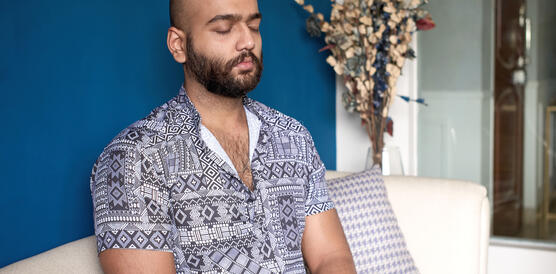 Ein indischer Mann sitzt auf einer Couch mit den Beinen im Schneidersitz und hat die Augen zur Meditation geschlossen.