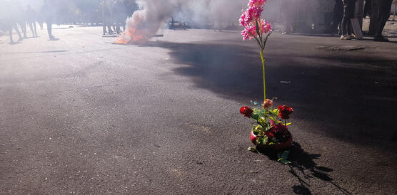 Auf einer Straße steigt Rauch von einem Feuer auf, Menschen stehen ringsum und mitten auf der Straße: ein Blumenbouqet.