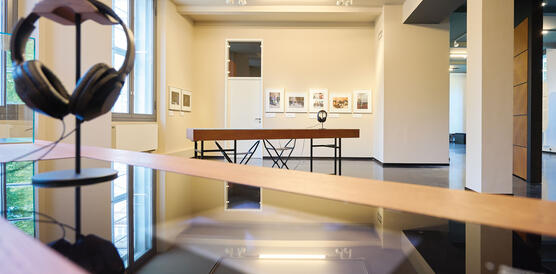 Ein Museumsraum, in dem Tische stehen, Bilder an der Wand hängen; auf einem Tisch ist ein Kopfhörer auf einem Bügel aufgehängt.