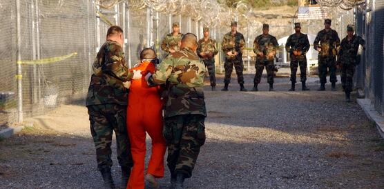 Das Bild zeigt Soldaten wie sie einen Mann in orangenem Overall abführen. Seitlich ist ein Zaun mit Stacheldraht zu sehen.