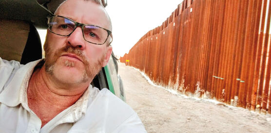 Ein Mann mit Bart, Brille und Hemd sitzt in einem Auto und fährt an einem Grenzzaun entlang.