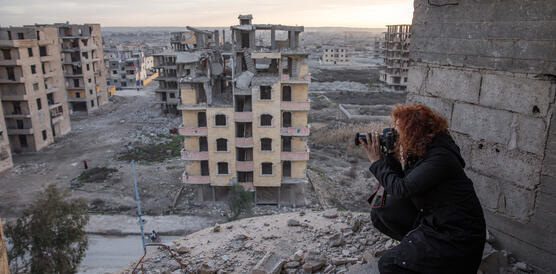 Das Bild zeigt eine Frau mit einer Fotokamera, die inmitten von Häuserruinen sitzt und ein Foto macht