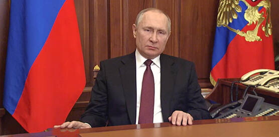 Das Bild zeigt Wladimir Putin, der einen Anzug mit Krawatte trägt und an einem Holztisch sitzt. Hinter ihm steht die russische Fahne.