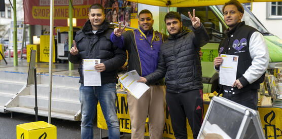 Das Bild zeigt vier junge Männer, die vor einem gelben Amnesty-Stand stehen und in die Kamera gucken. Manche zeigen ein Peace-Zeichen und halten einen Brief vom Briefmarathon in den Händen.