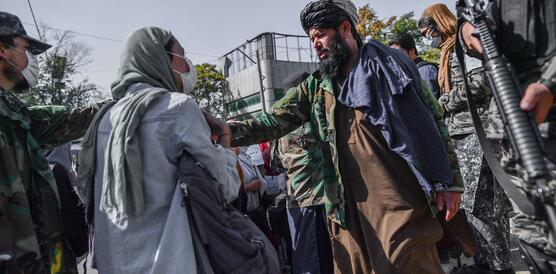 Eine Frau mit Kopftuch steht vor einem Taliban, der sie mit der rechten Hand leicht wegschiebt. Um die beiden Personen herum stehen weitere bewaffnete Taliban.