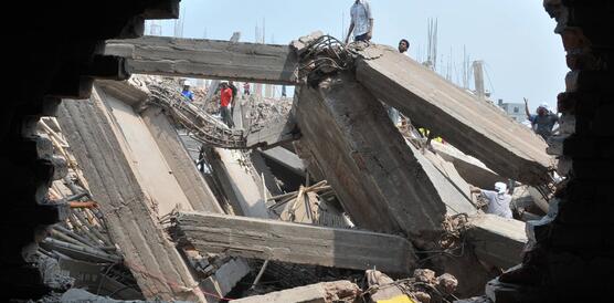 Das Bild zeigt die Trümmer eines eingestürzten Gebäudes, in den Trümmern stehen Menschen