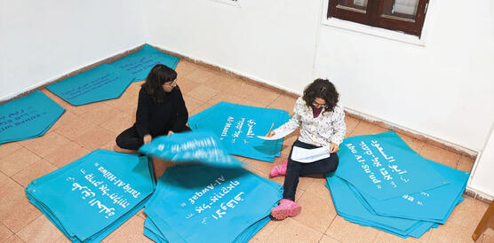 Zwei Frauen sitzen auf dem Boden und arbeiten an Bannern aus Plane, auf die arabische Straßennamen gedruckt sind.
