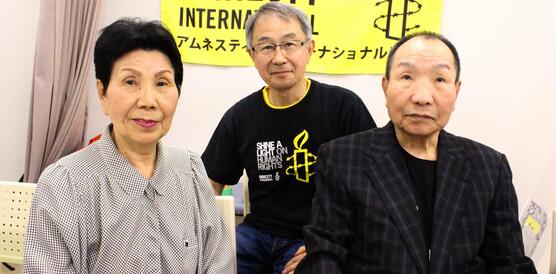 Drei Personen sitzen nebeneinander in einem Zimmer. An der Wand hinter ihnen hängt ein Banner mit der Aufschrift "Amnesty International".