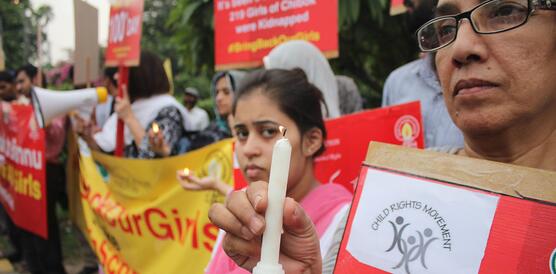Eine Frau hält in der rechten Hand eine Kerze und in der linken Hand ein Plakat. Sie steht vor einer größeren Gruppe Menschen, die ebenfalls Kerzen und Plakate und Banner halten.