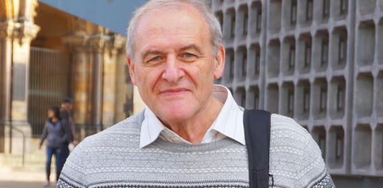 Ein älterer Mann, graues Haar, lächelnd, in Hemd und Pullover, Rucksack über die linke Schulter hängend, draußen vor einem Gebäude.