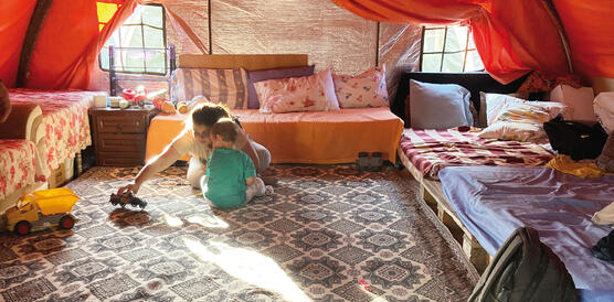 Ein Zelt in einem Zeltlager, der Boden ist mit Pappe und Teppichen ausgelegt, an den Rändern stehen improvisierte Sofas, zwei Kinder spielen auf dem Boden mit Spielzeugautos.