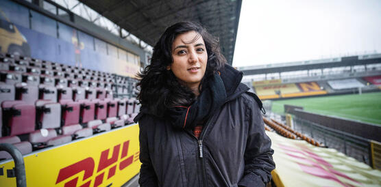 Eine junge Frau mit gewelltem Haar trägt eine Jacke und einen Schal und steht auf der Tribüne eines Fußballstadions.