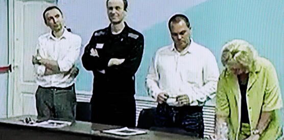 Vier Personen stehen nebeneinander hinter einem Tisch in einem Gerichtssaal. Nawalny hat die Arme verschränkt.