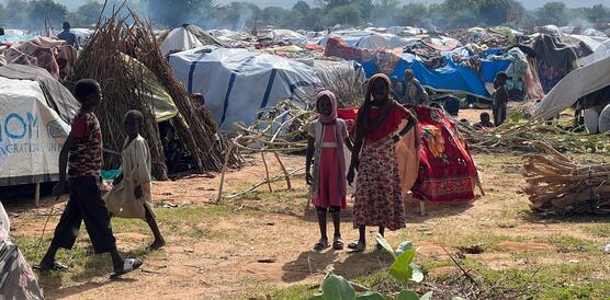 Ein paar Menschen, die wohl aus dem Sudan geflüchtet sind, inmitten einer Zeltstadt. Die sogenannten Zelte bestehen teilweise aus Plastikplanen, teilweise aus Ästen. 