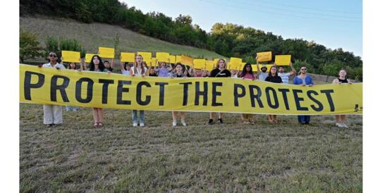 Eine Gruppe von jungen Menschen steht auf einer Wiese, die vorderen halten ein sehr langes großes gelbes Banner mit der schwarzen Aufschrift "Protect the Protest" vor sich, die anderen dahinter halten kleine gelber Schilder mit weiteren Aufschriften hoch. 