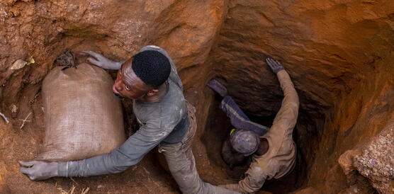 Zwei Männer entsteigen einem Erdloch, das in eine Mine führt, einer von ihnen hat einen prallgefüllten Stoffsackin den Armen.
