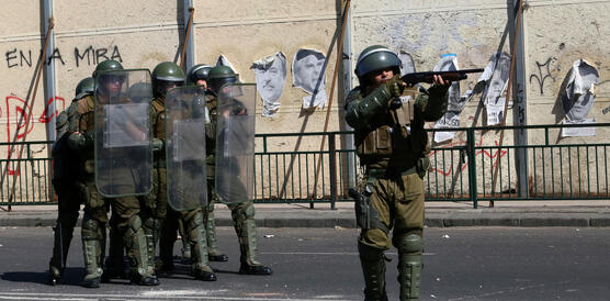 Das Foto zeigt eine Gruppe von sechs Polizisten mit Helmen und Schutzschildern. Vor ihnen steht ein weiterer Polizist mit Helm, der mit einem Gewehr anlegt und zielt.