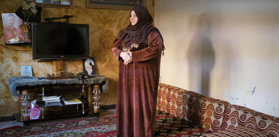 Eine muslimische Frau in traditionellem Gewand und mit Kopftuch steht in ihrem WOhmnzimmer auf einem Teppichboden, rechts von ihr ein Fernseher und Bilder an der Wand; ihre Arme hält sie auf Bauchhöhe vor ihrem Körper, die Hände berühren sich dabei.