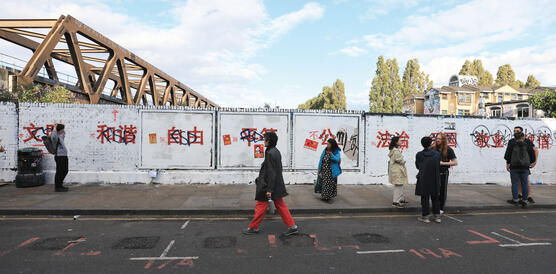 Eine lange Ziegelmauer, die mit weißer Farbe übermalt wurde, darauf in rot chinesische Schriftzeichen, Passant*innen gehen davor entlang.