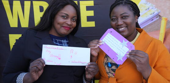 Das Bild zeigt zwei Schwarze Frauen, die in die Kamera schauen, lächeln und dabei jeweils selbstgeschriebene, bunte Briefe in die Kamera halten.